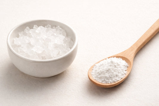 Ceramic bowl of alum crystals with a wooden spoon of alum powder on a neutral background, showing both forms of alum for natural dyeing.