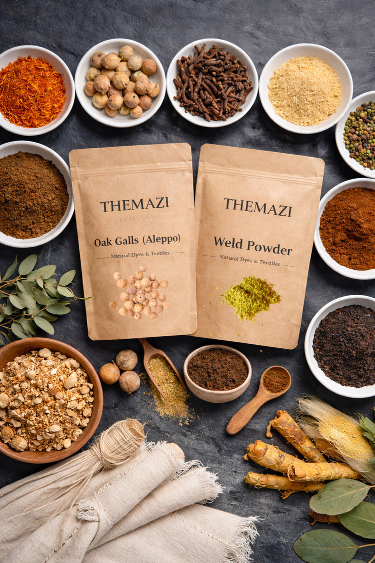 Natural dye ingredients and mordants arranged on a dark slate surface, with kraft paper packaging and plant materials.