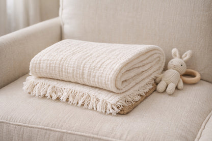 Undyed 4-layer crinkle muslin baby blanket with fringed edges, folded on a neutral nursing chair (115x115 cm / 45x45 in).
